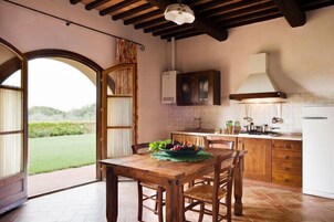 Cozinha rústica com armários de madeira, uma mesa de jantar com cadeiras e vista para uma paisagem verde através de uma abertura.