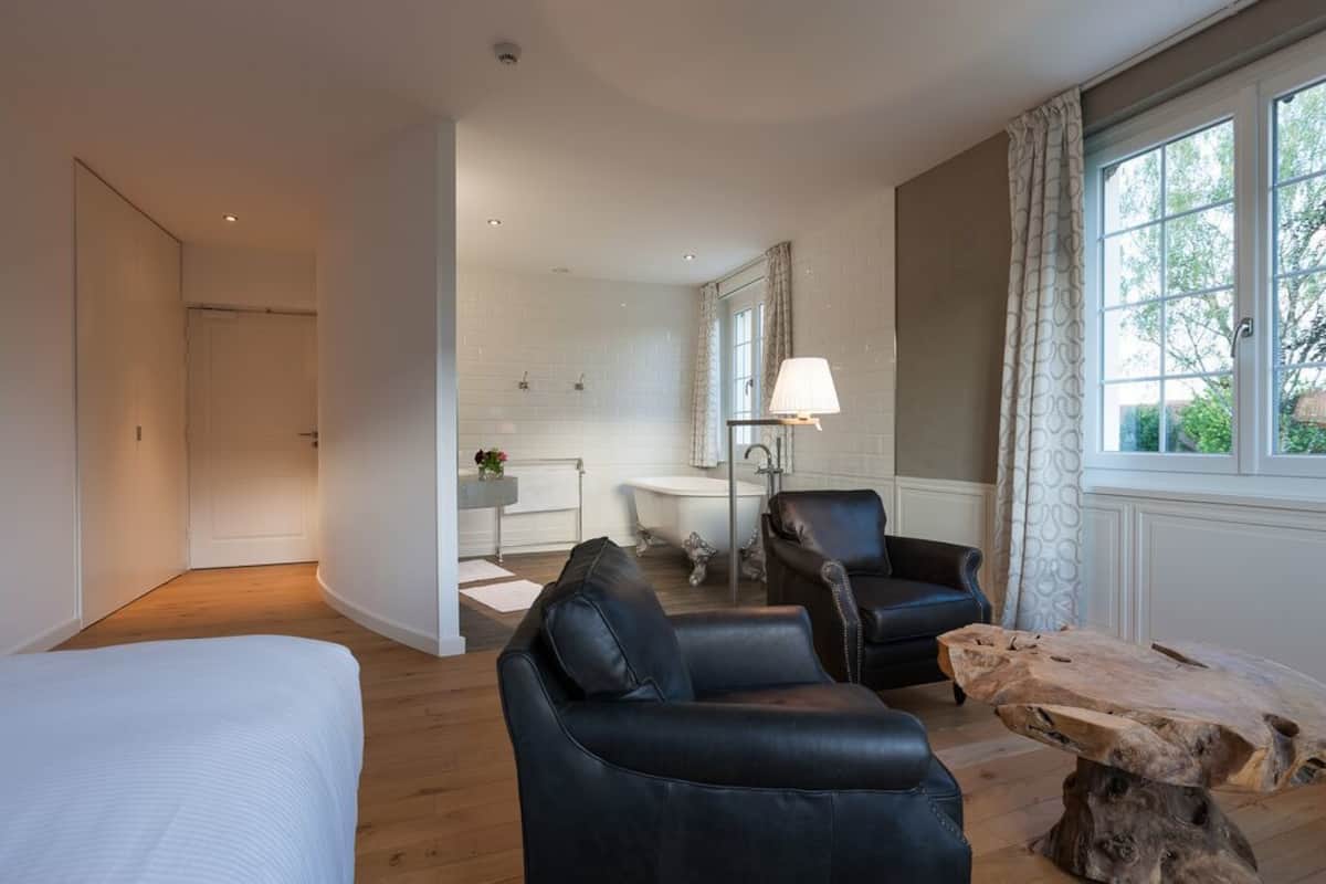 Un hotel moderno con una cama grande, sillones de cuero, una mesa de centro de madera, un baño con bañera y vistas a zonas verdes por la ventana.