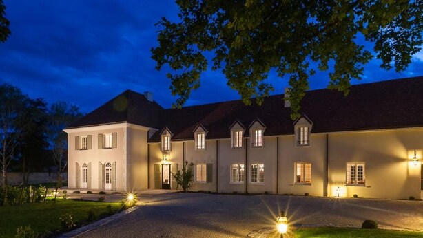 A large, multi-story house with a driveway leading to the entrance, surrounded by trees and well-lit exterior lighting.
