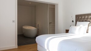 A bedroom with a large bed, a wooden floor, a white door leading to a bathroom with a bathtub, and a nightstand with a lamp.