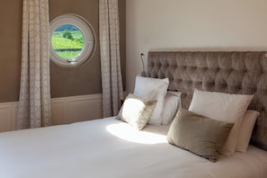 A bed with a tufted headboard, a window with a view of a green field, and patterned curtains.