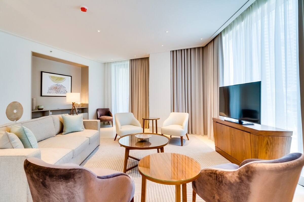 A modern hotel room with a sofa, armchairs, a coffee table, a TV, and a wooden cabinet.