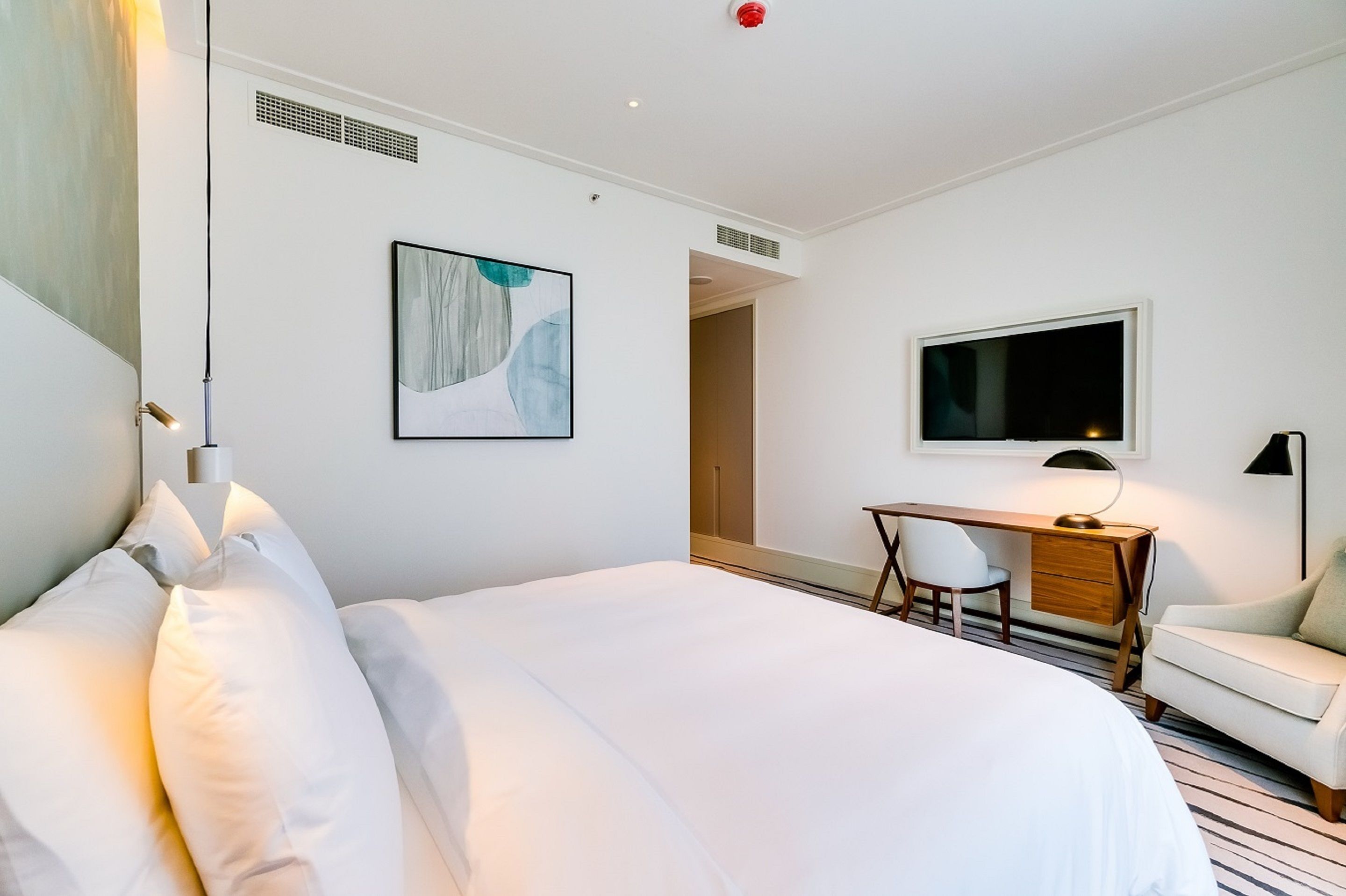Quarto de hotel com uma cama grande, uma escrivaninha com cadeira, uma televisão e um quadro na parede.