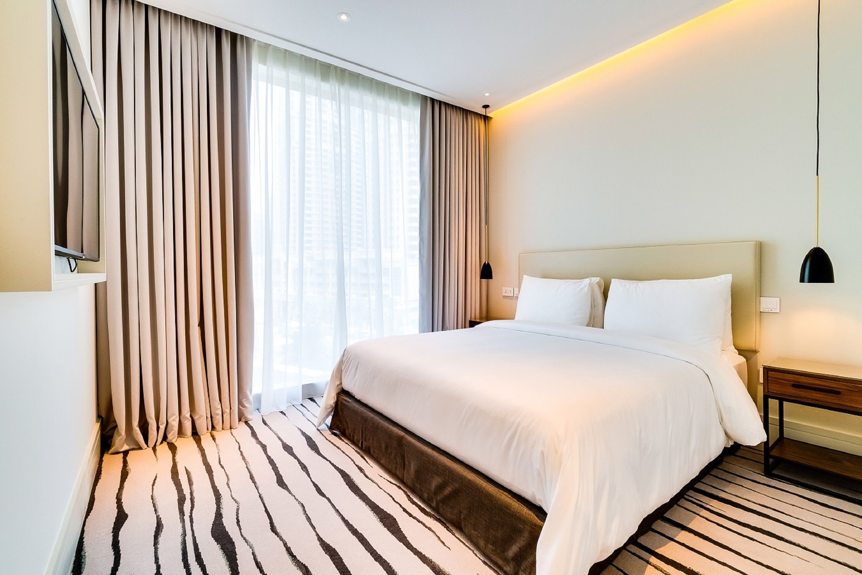 A hotel room with a large bed, a flat-screen TV mounted on the wall, a bedside table with a lamp, and a striped carpet.