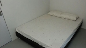 Um quarto pequeno, com uma cama de solteiro, colchão branco simples e cabeceira sem adornos.