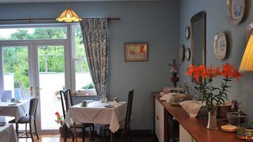 Un comedor con una mesa lista para dos, una ventana con cortinas y vistas al exterior con vegetación.