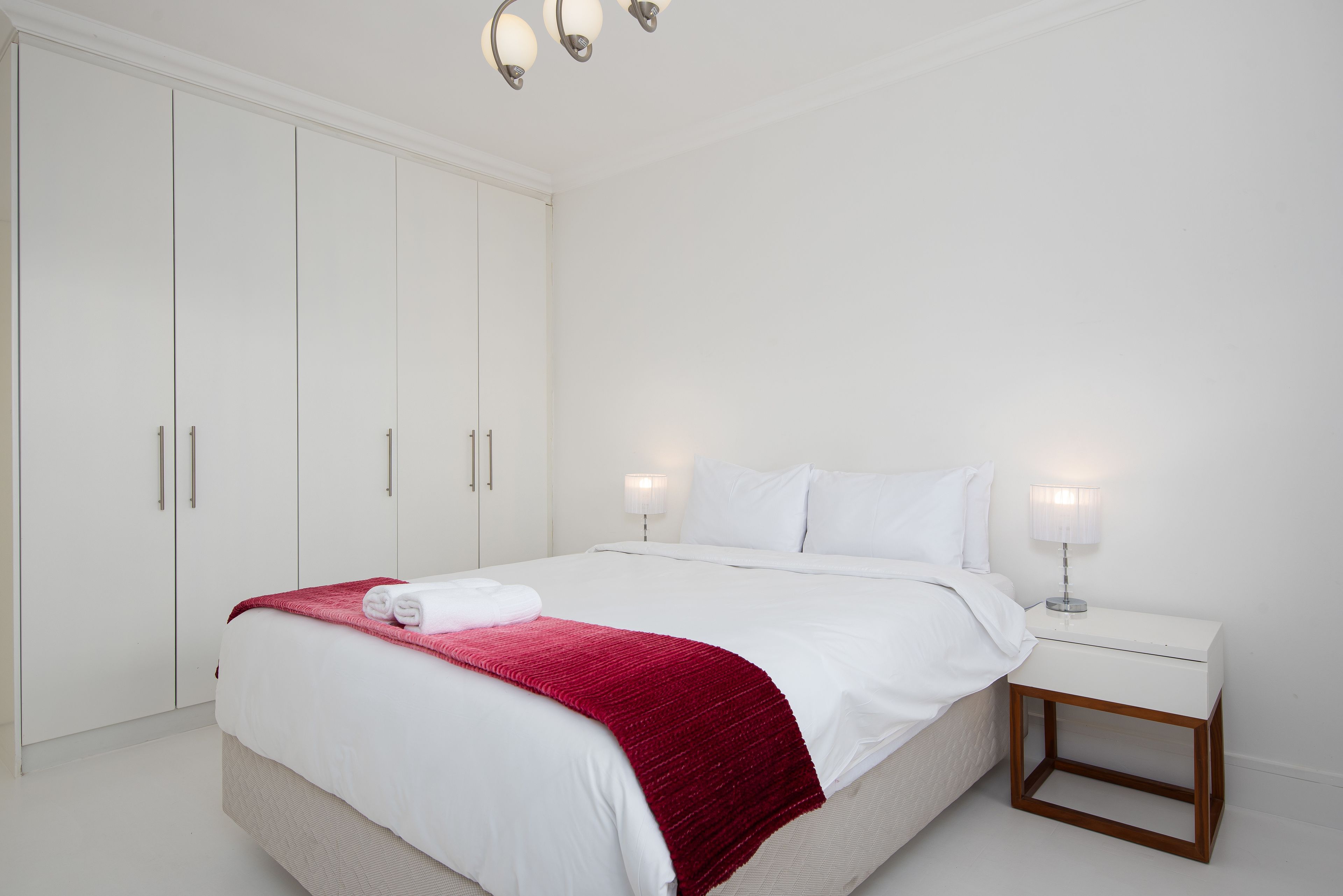 A neatly made bed with white linens and a red blanket, a white bedside table with a lamp, and a wardrobe with silver handles.