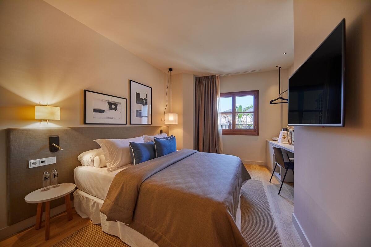 A hotel room with a bed, bedside table, lamp, artwork, and a desk with a chair.