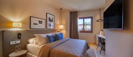 A hotel room with a bed, bedside table, lamp, artwork, and a desk with a chair.