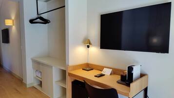 Habitación de hotel con escritorio de madera, silla negra, televisor montado en la pared, perchero y lámpara de noche.
