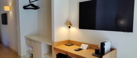 Een hotelkamer met een houten bureau, een zwarte stoel, een aan de muur bevestigde televisie, een kapstok en een bedlampje.