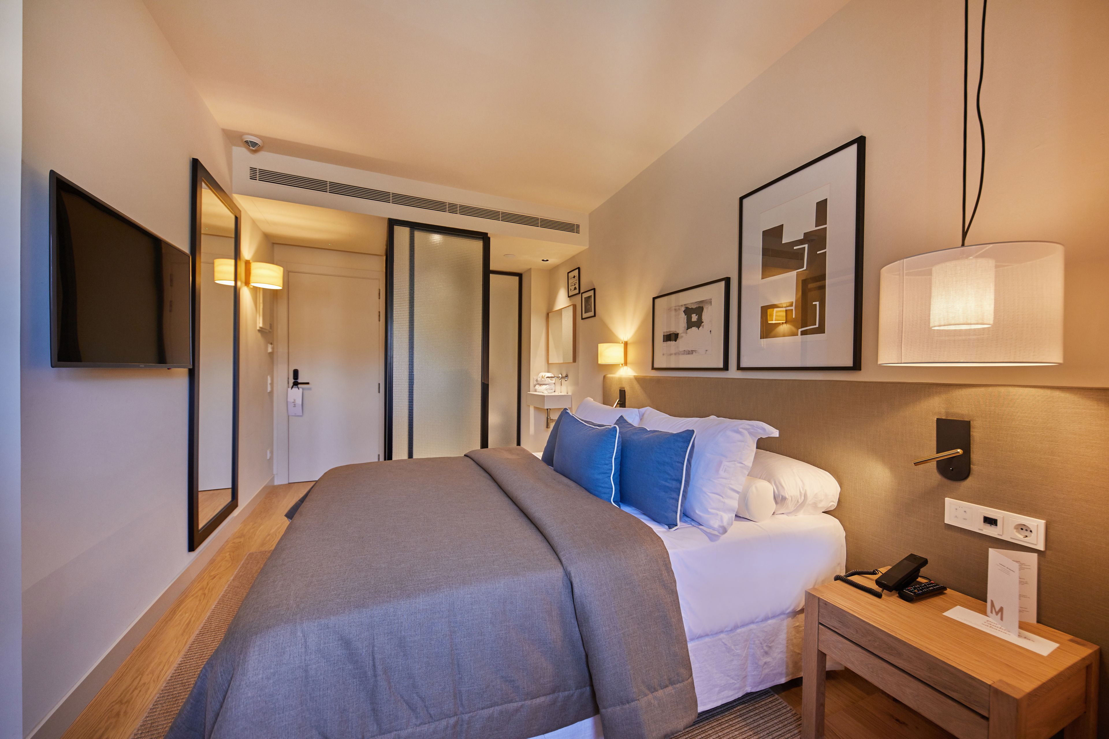 Una habitación de hotel con cama, mesita de noche, lámpara y televisor montado en la pared.