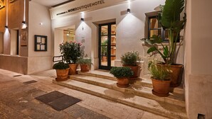 The entrance to MonBoutiqueHotel with a sign above the door, potted plants in front, and a well-lit interior visible through the glass doors. - Mon Boutique Hotel (Pollenca)