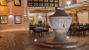 An outdoor seating area with stone tables and chairs, a large stone fountain, and umbrellas providing shade. - Mon Boutique Hotel (Pollenca)