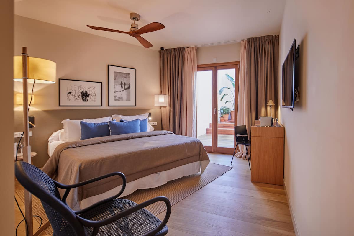 A hotel room with a bed, a desk, a chair, a ceiling fan, and a TV.