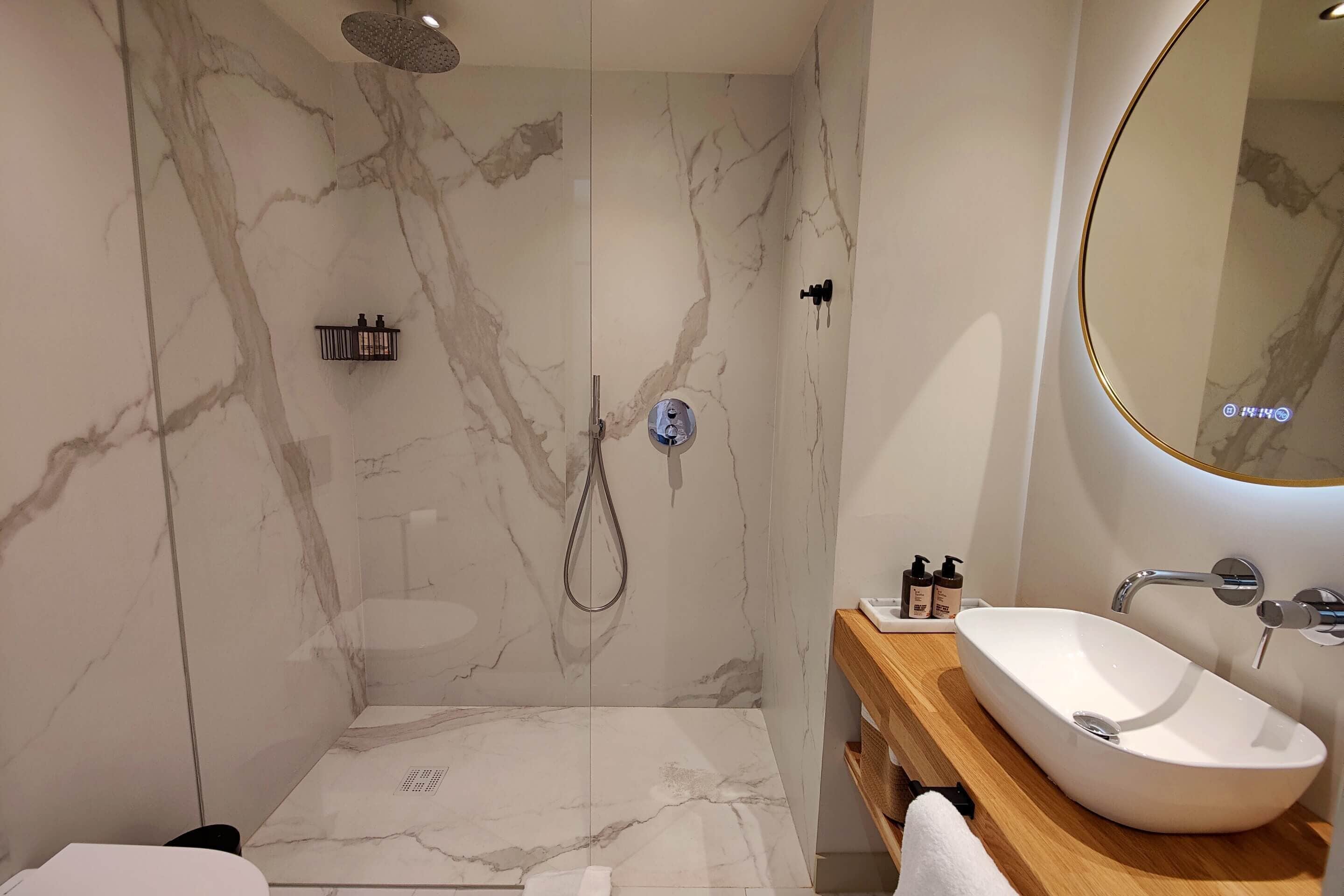 Un baño moderno con pared de ducha de mármol, una bañera independiente y un tocador de madera con lavamanos.