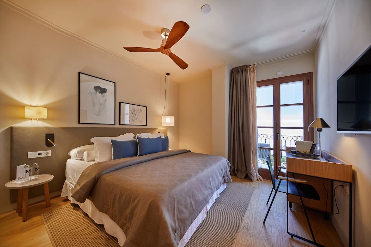 A hotel room with a bed, desk, ceiling fan, artwork, and a balcony view.