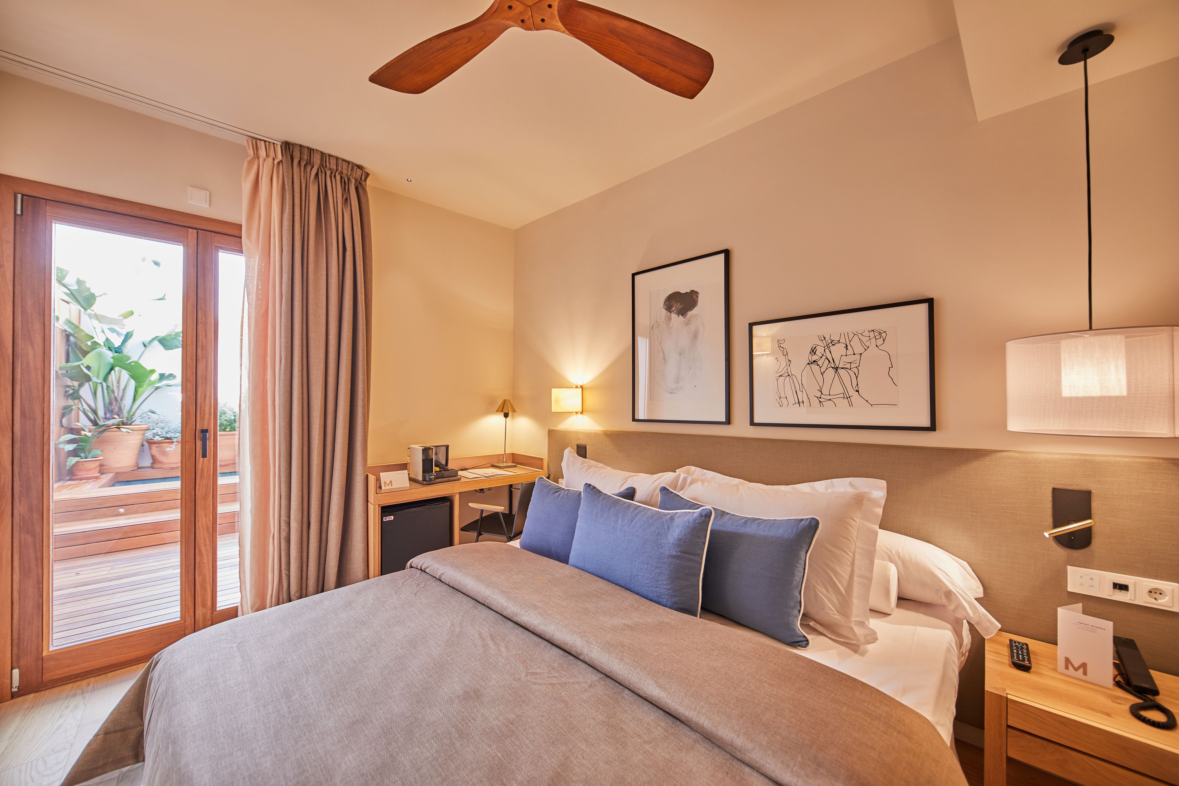 A hotel room with a bed, a desk, a lamp, a ceiling fan, and a view of the outdoors.