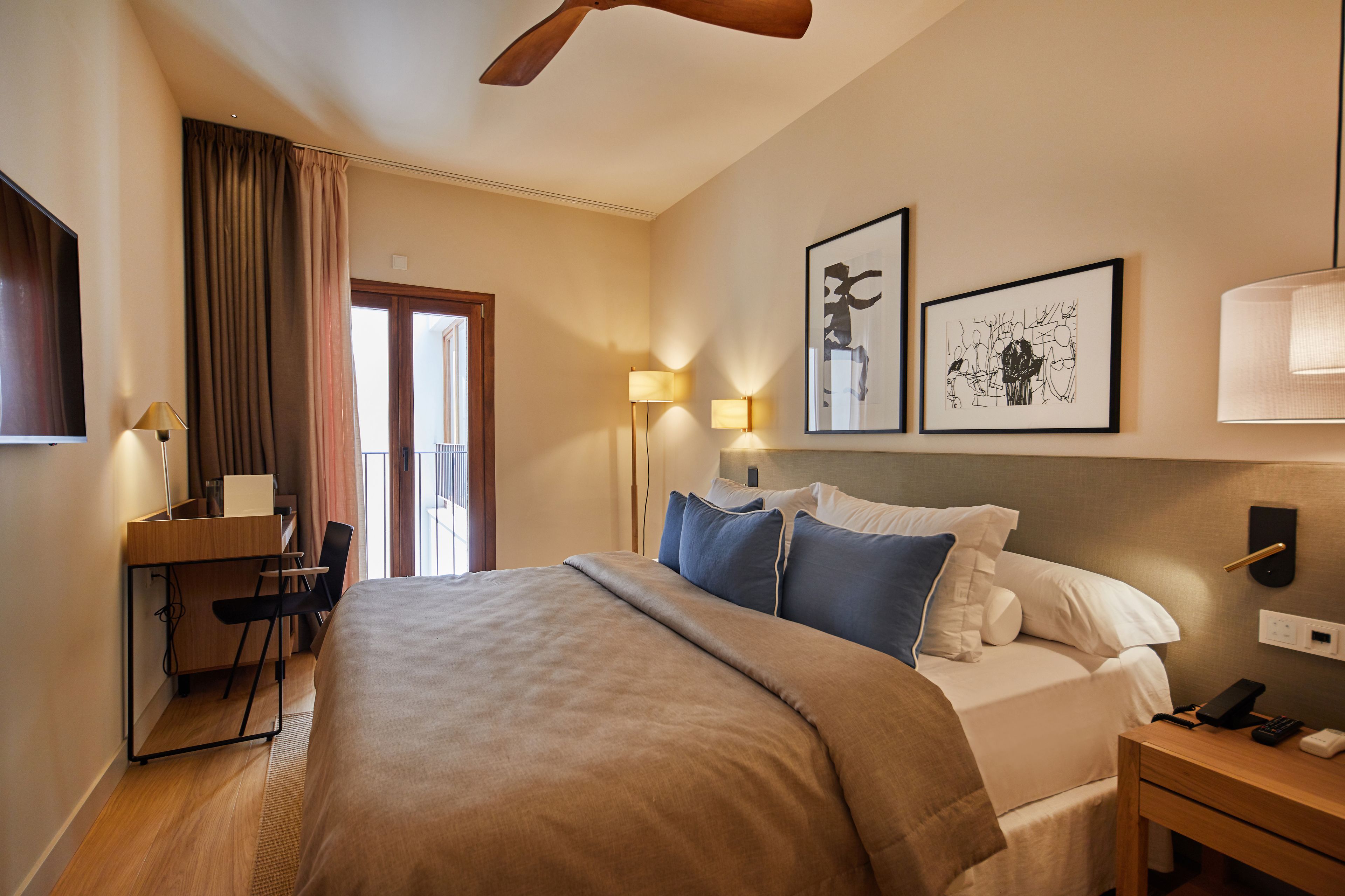 A hotel room with a bed, desk, chair, lamp, artwork, and a ceiling fan.
