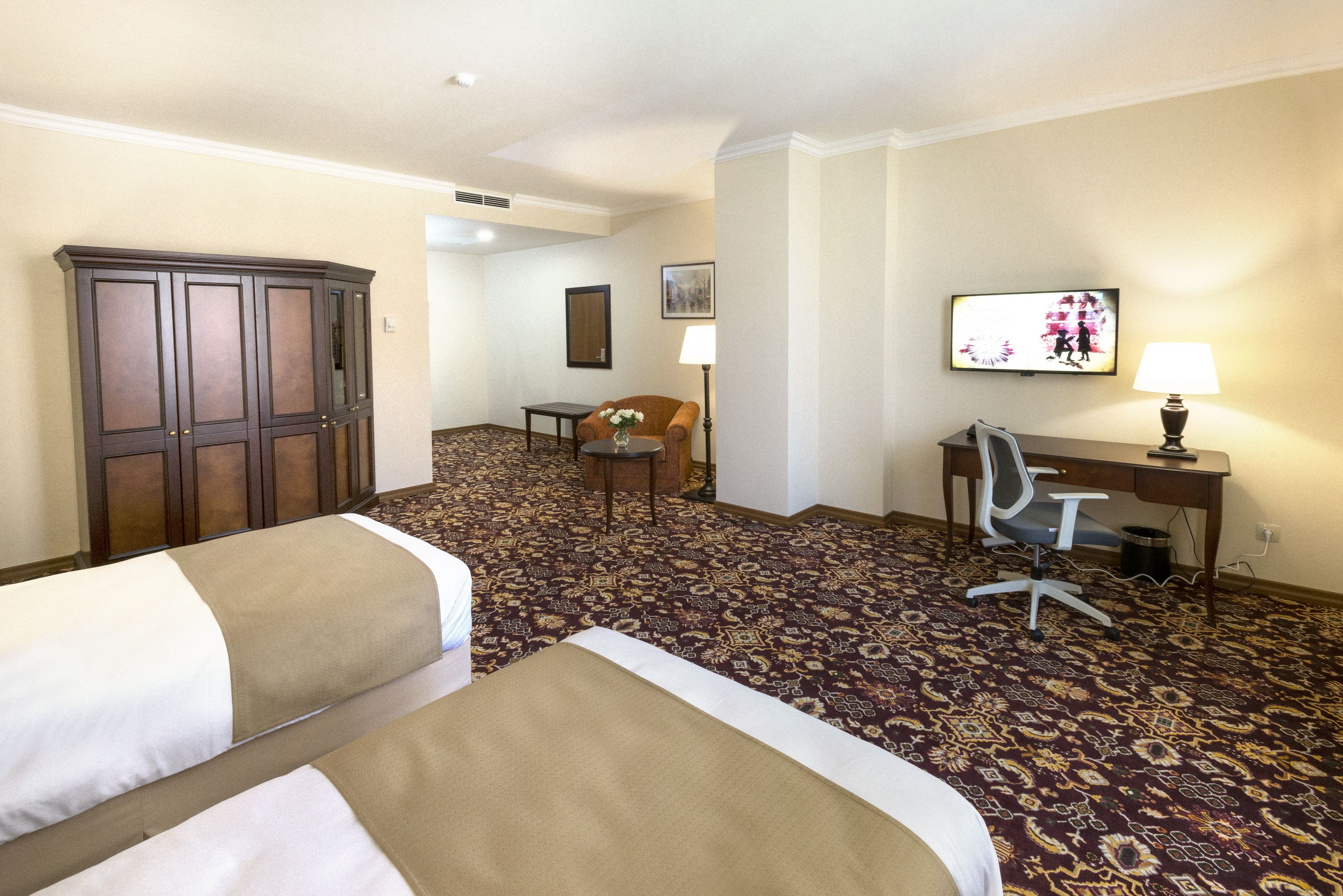 A hotel room with two beds, a desk, a chair, a lamp, and a television.