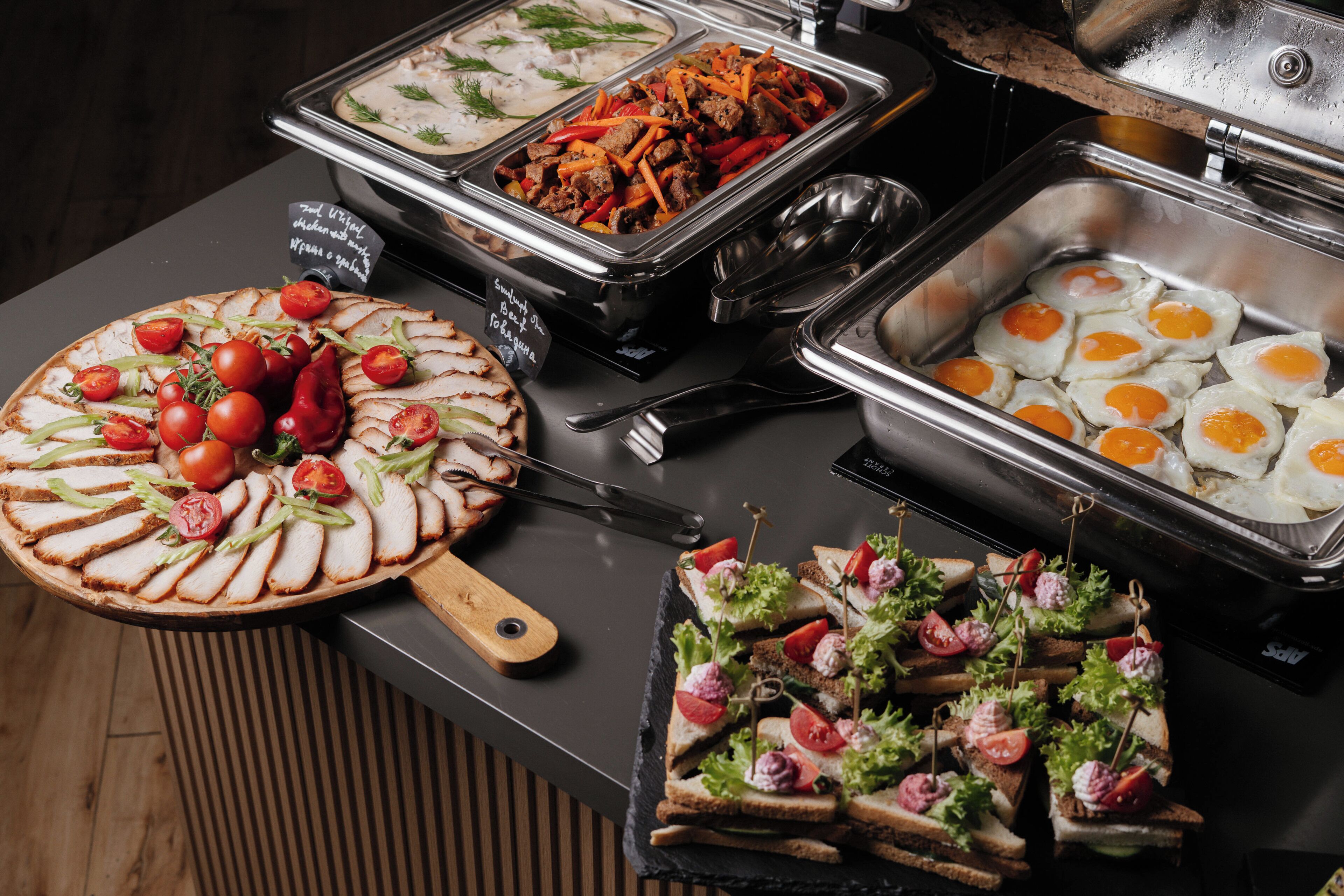 Un bufé con diversas opciones de comida, incluyendo una bandeja de carnes frías, platos de huevo y sándwiches.
