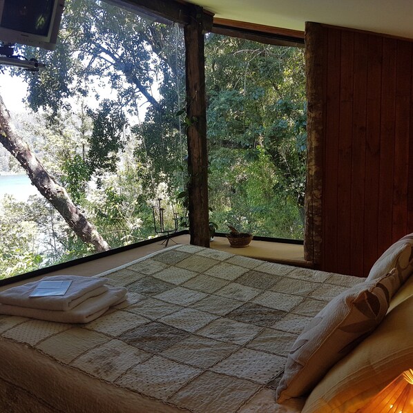 A bedroom with a large window, a bed, and a view of trees and a body of water.