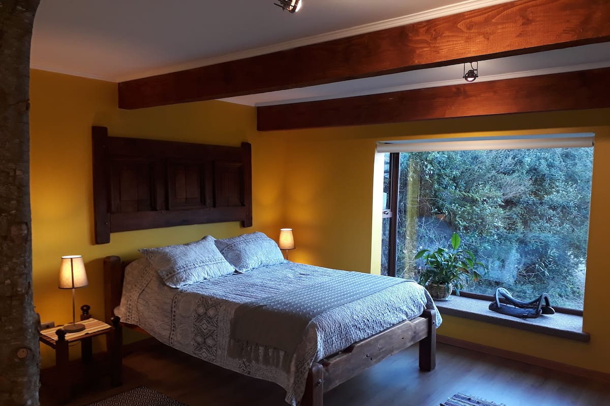 A bedroom with a wooden bed, two bedside lamps, a patterned rug, and a window with a view of greenery.