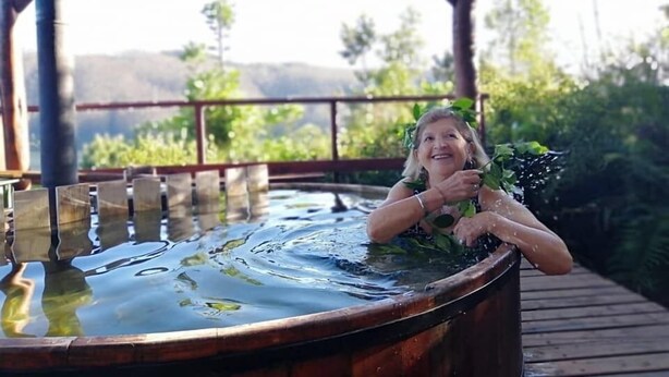 A person relaxing in a wooden hot tub with a view of greenery and hills in the background.