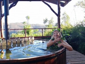 A person relaxing in a wooden hot tub with a view of greenery and hills in the background.