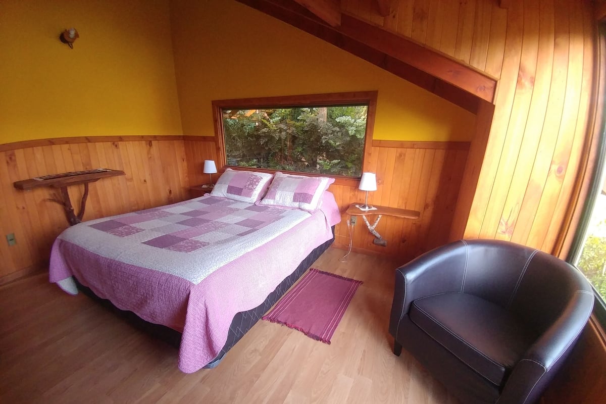 A cozy bedroom with a wooden bed, a chair, and a window with a view of greenery.