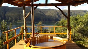 A wooden deck with a hot tub, surrounded by a wooden railing and a pergola, overlooking a lake and forested hills.