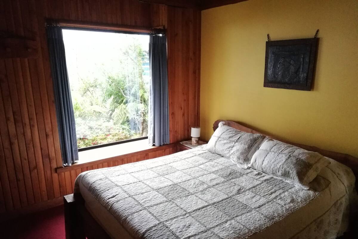A bedroom with a wooden floor, a bed with a quilt, a nightstand with a lamp, a window with curtains, and a wall-mounted picture.