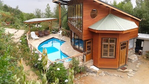 A wooden cabin with a pool, surrounded by lush greenery and a lake view.