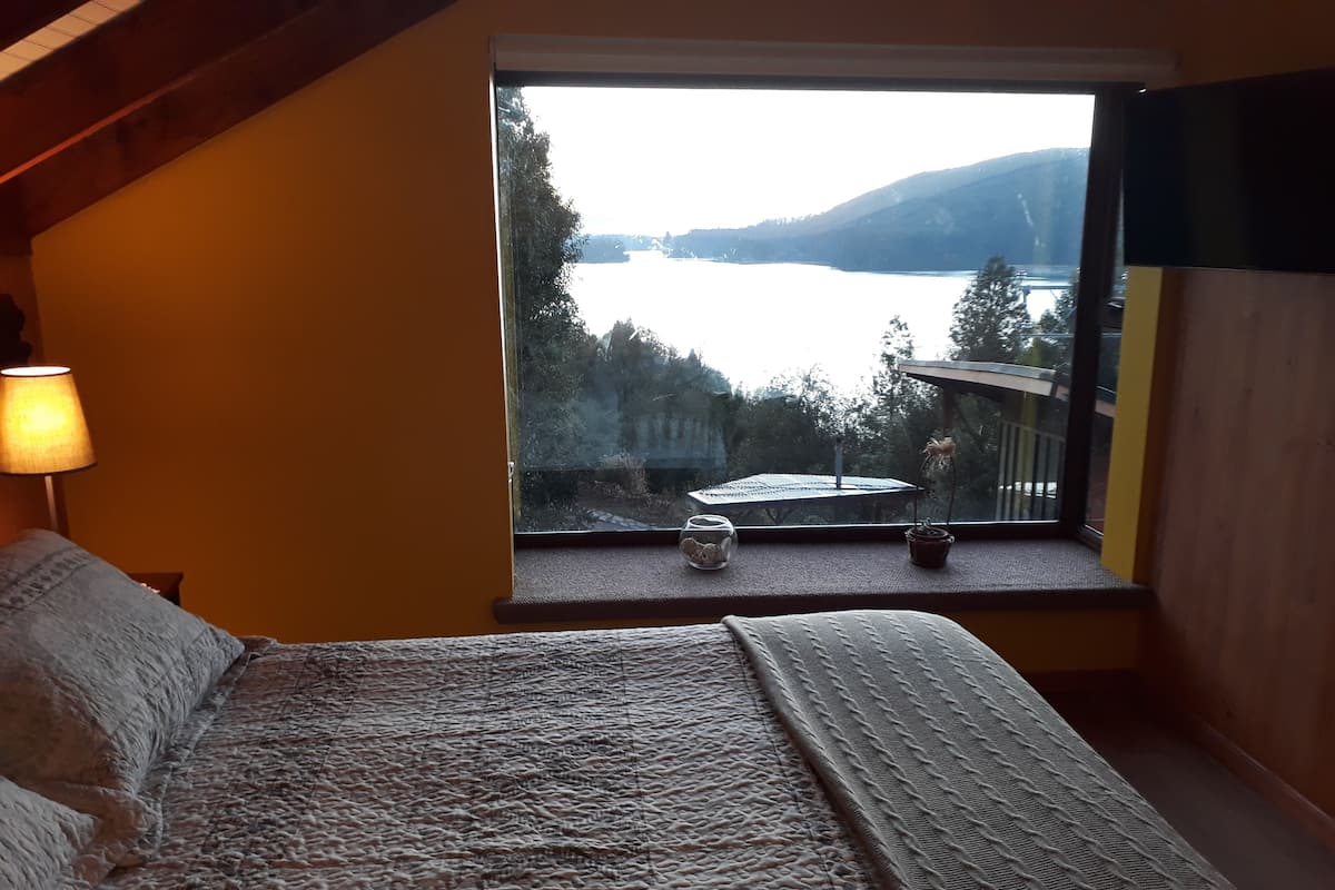 A bedroom with a large window overlooking a lake and forest. There is a bed with a textured blanket, a lamp, and a small table with a plant and a decorative item.