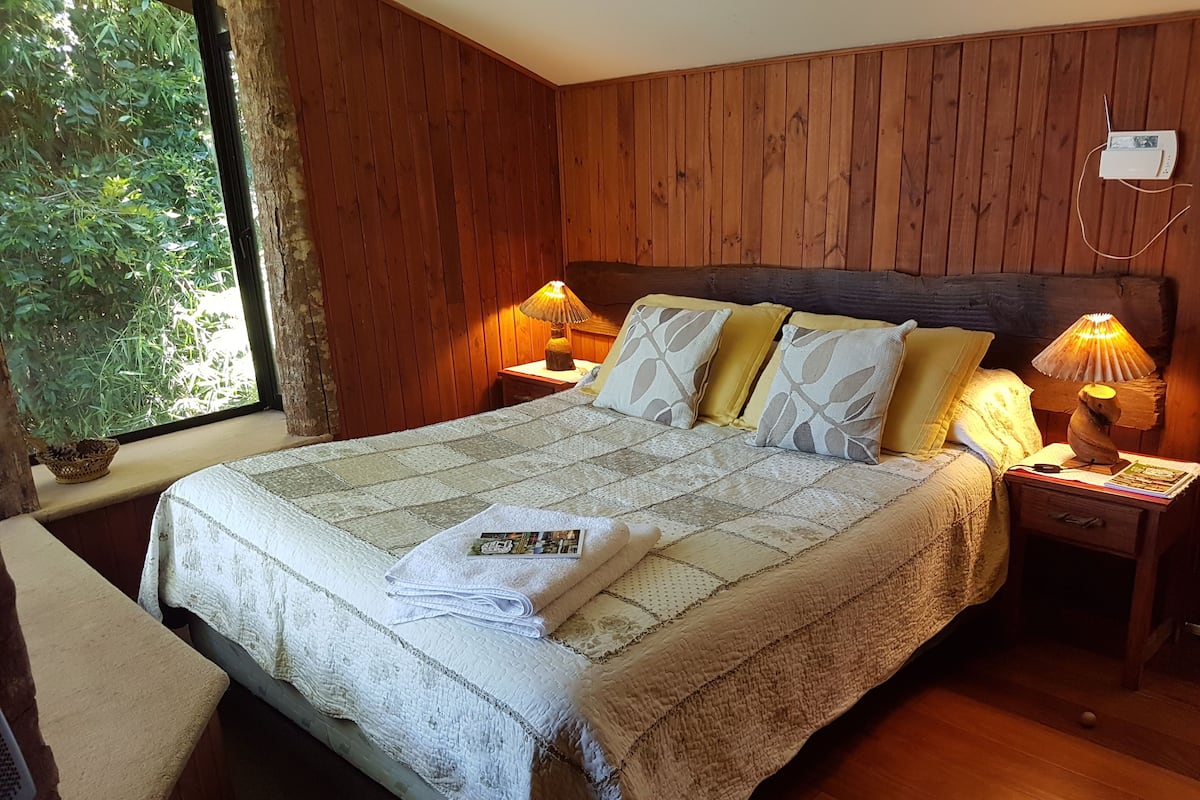 A neatly made bed with a quilt, pillows, and a folded towel. There are two bedside lamps and a small table with a lamp. A window with a view of greenery is visible.