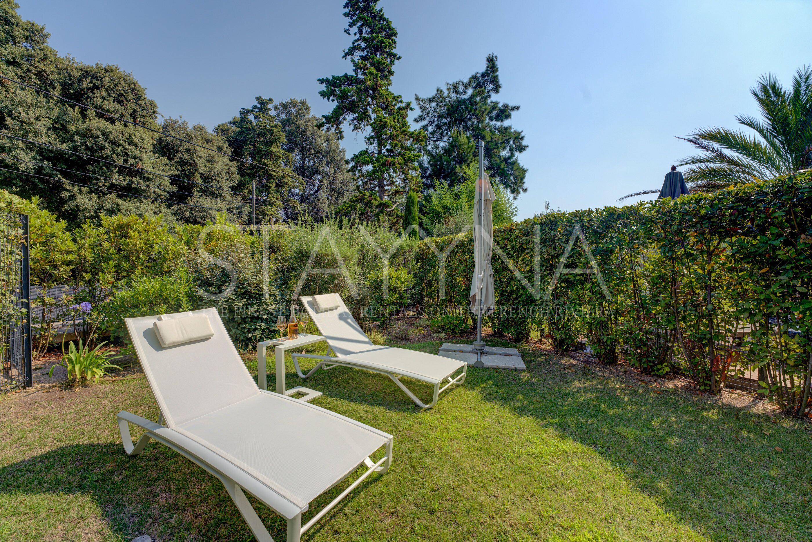 Un jardín con dos sillones blancos y una pequeña mesa, rodeado de follaje y árboles.