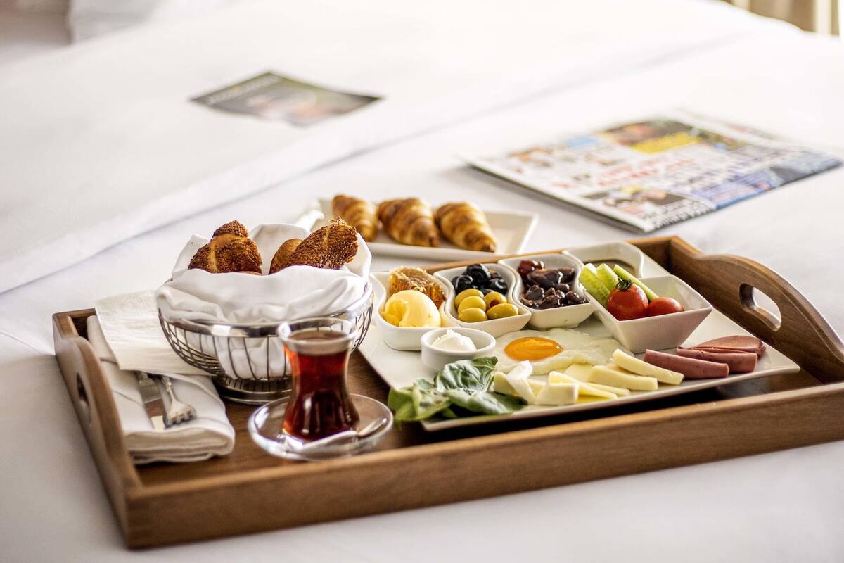 En bakke med morgenmad, blandt andet croissanter, ost, æg og forskelligt frugt, placeret på en seng.