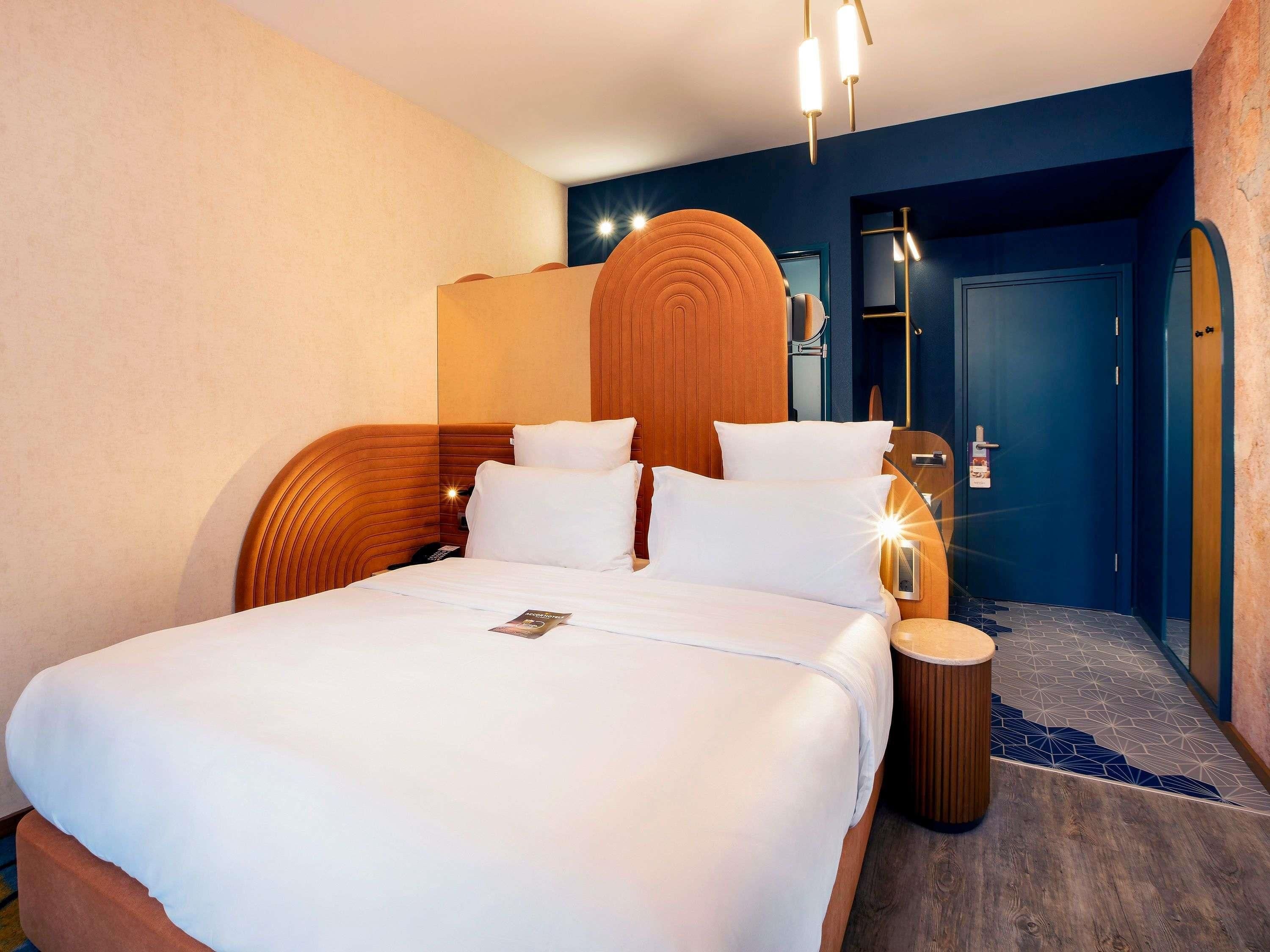 A hotel room with a large bed, a wooden headboard, a round wall mirror, and a blue door.