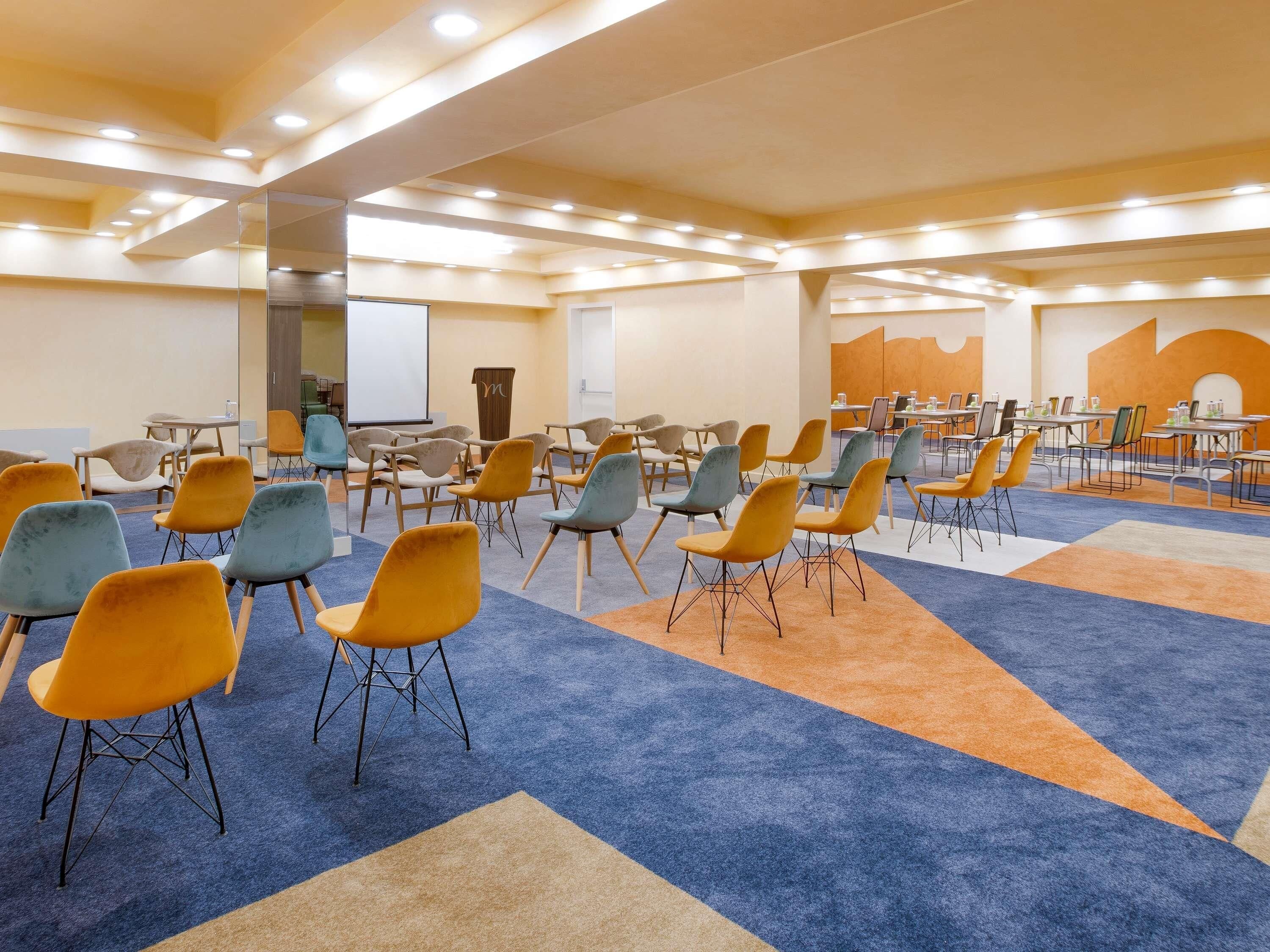 Une salle de conférence avec des rangées de chaises, des tables et un écran de projection.