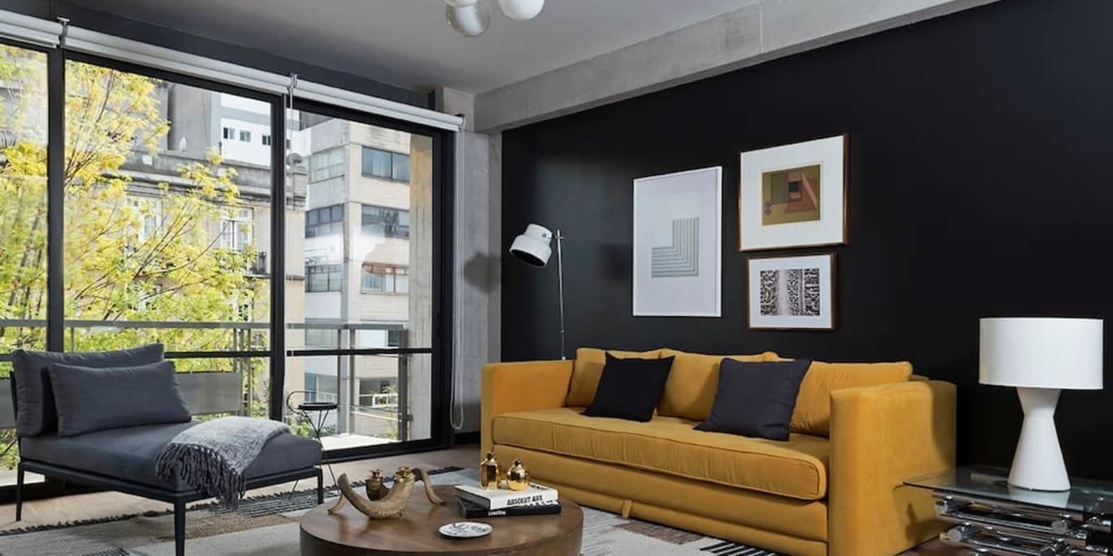 A modern living room with a sofa, coffee table, and a lamp. There are large windows providing a view of buildings and trees.