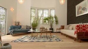 A modern living room with a blue sofa, a white sofa with a red and white checkered throw, a coffee table, and a patterned rug. There are large windows with a view of greenery outside. - Unique Roma Town House by Mr.W (Mexico City)