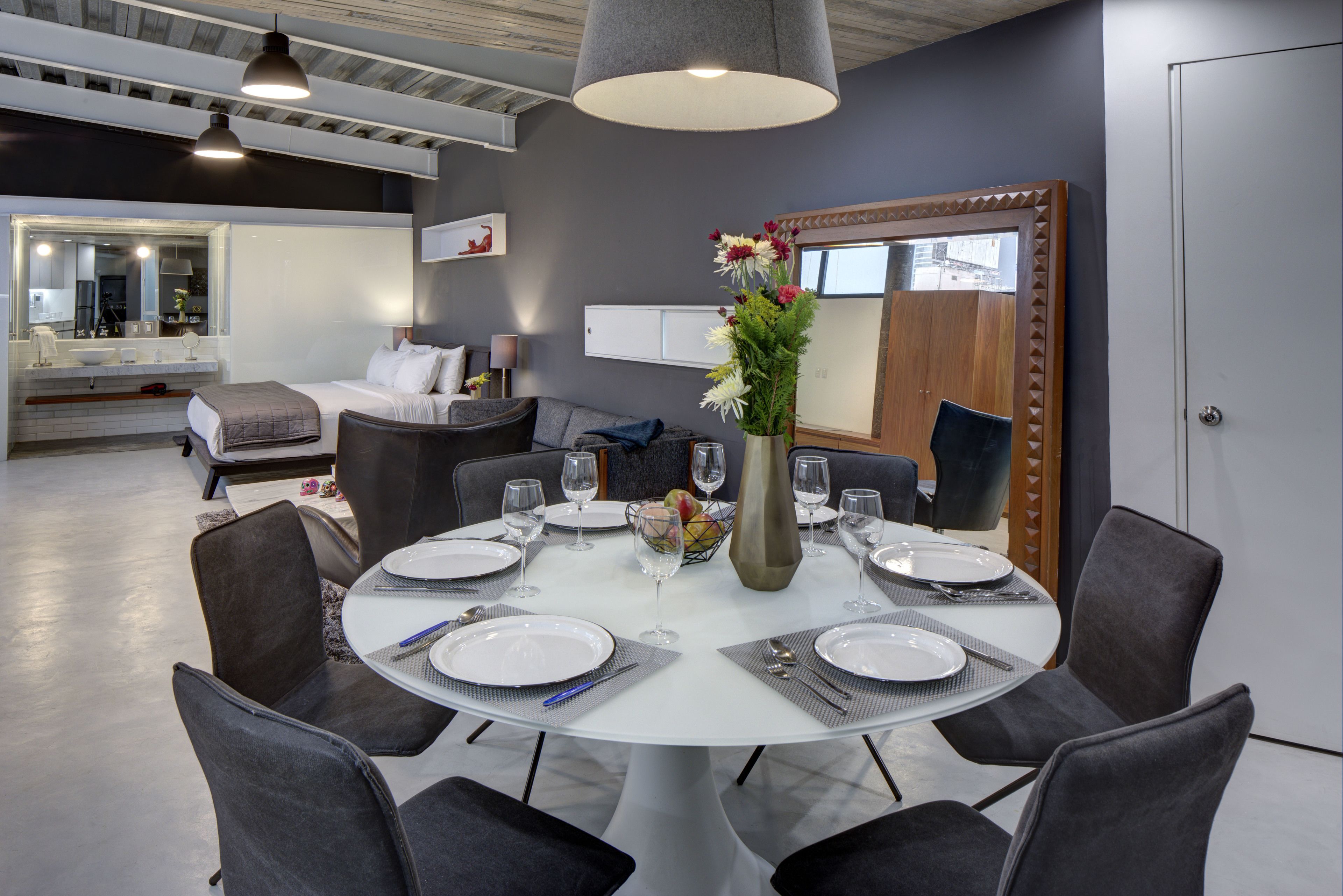 Een eetruimte met een ronde tafel gedekt voor zes personen, omringd door zes stoelen. Op tafel staat een vaas met bloemen en de kamer heeft een modern design met grijze wanden en een plafond met zichtbare balken.
