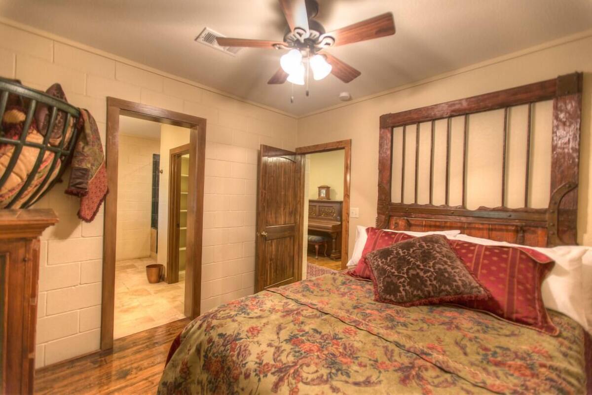 Une chambre à coucher avec un lit, une tête de lit en bois, un ventilateur de plafond et une porte donnant sur une autre pièce.