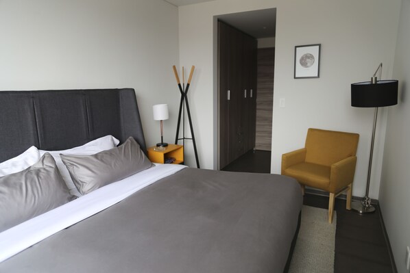 Une chambre d’hôtel moderne avec un grand lit, une petite table de chevet, une chaise, une lampe sur pied et un tableau encadré au mur.