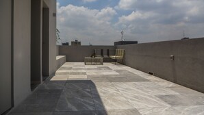 Une terrasse sur le toit, dotée d’un sol carrelé, d’un espace salon avec une table et des chaises, et offrant une vue sur les toits de la ville sous un ciel partiellement nuageux.