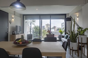 Un espace repas moderne avec une grande table, des chaises et un saladier rempli de fruits. Il y a un balcon agrémenté de plantes en pot et offrant une vue sur la ville.