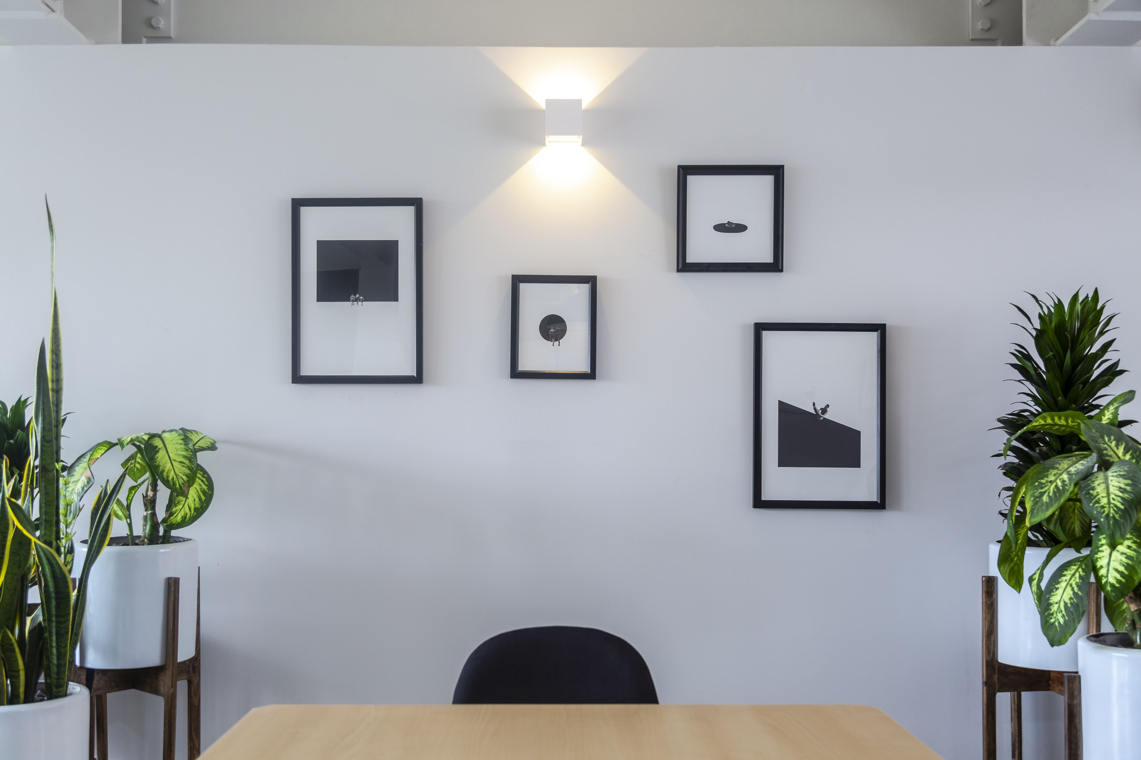A modern office space with a wooden table, four framed pictures on the wall, and potted plants on either side.