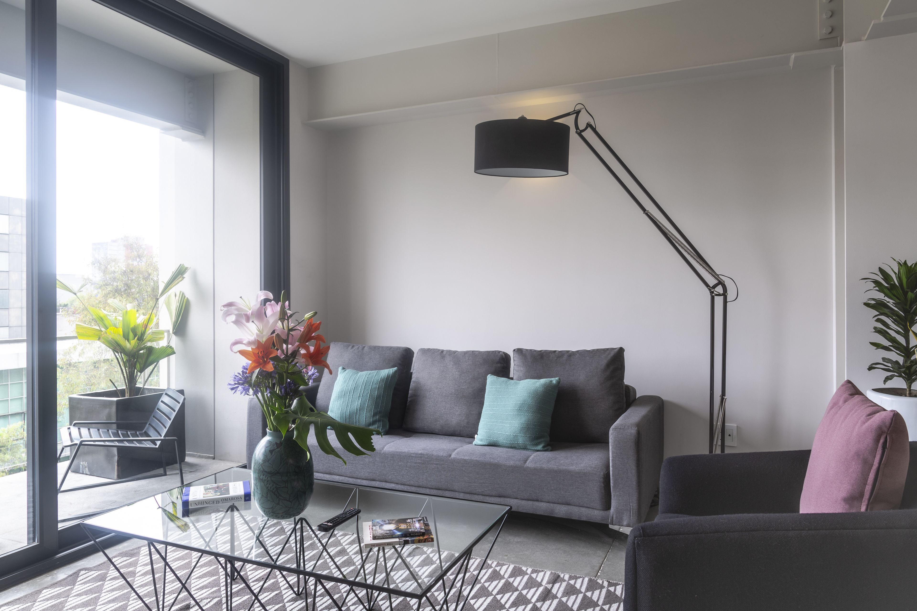 A modern living room with a grey sofa, a coffee table, and a floor lamp. There is a large window providing natural light and a view of greenery outside.