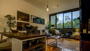 A modern living room with a wooden floor, a coffee table, a sofa, and a dining area with a wooden table and chairs. There are plants, a large window, and artwork on the walls. - Zen Modern by Mr.W (Mexico City)