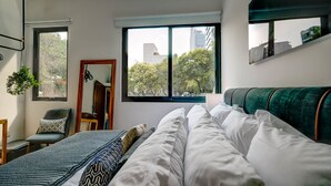 A modern bedroom with a large window, a bed with white and patterned bedding, a green upholstered headboard, a wooden chair, and a hanging plant. - Zen Modern by Mr.W (Mexico City)